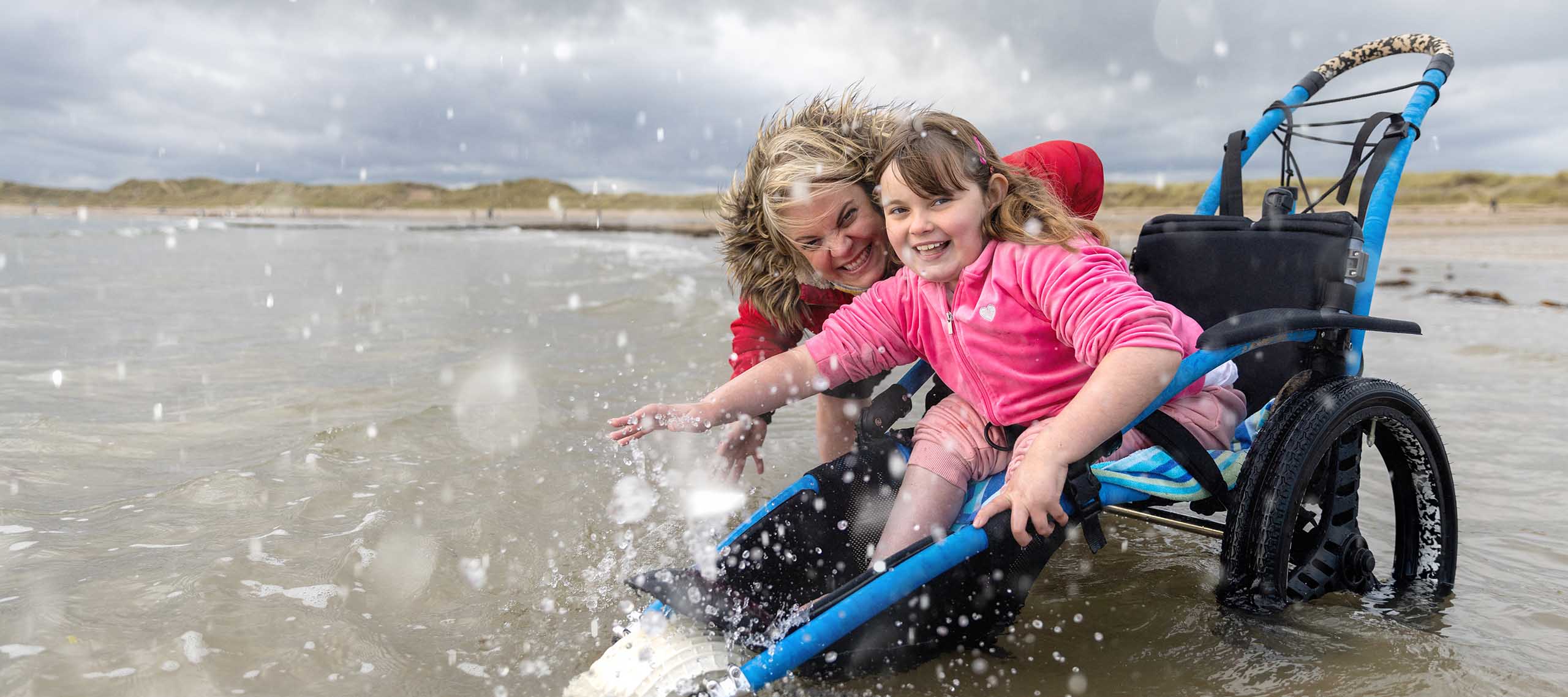 Mother and daughter in the sea in Beadnell, England, splashing around and smiling. The daughter is a wheelchair user.